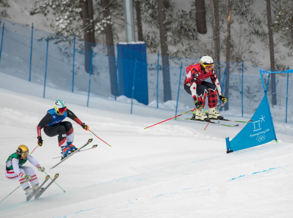 A ski cross racer dressed in red flies over a bump ahead of two other racers