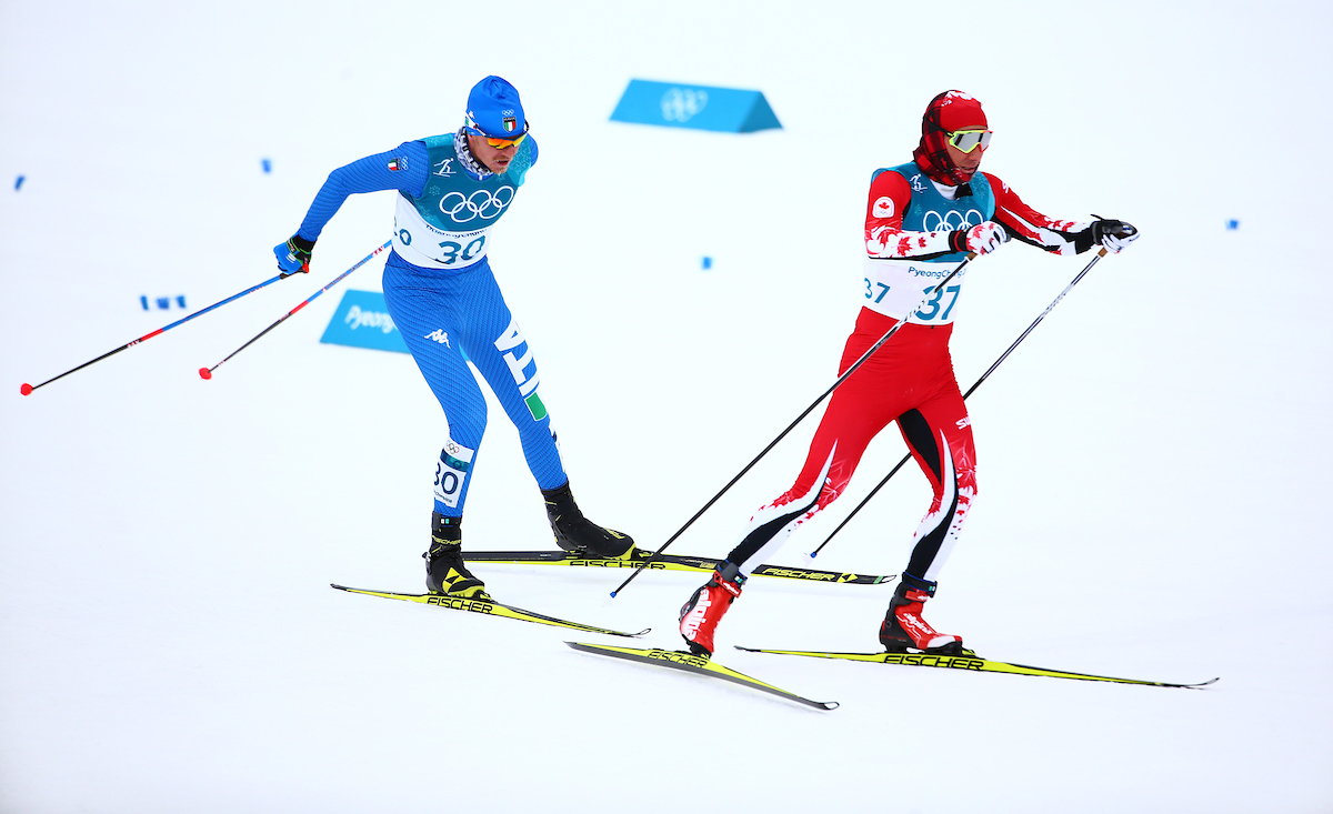 A cross-country skier in a red suit skis in front of a skier in blue
